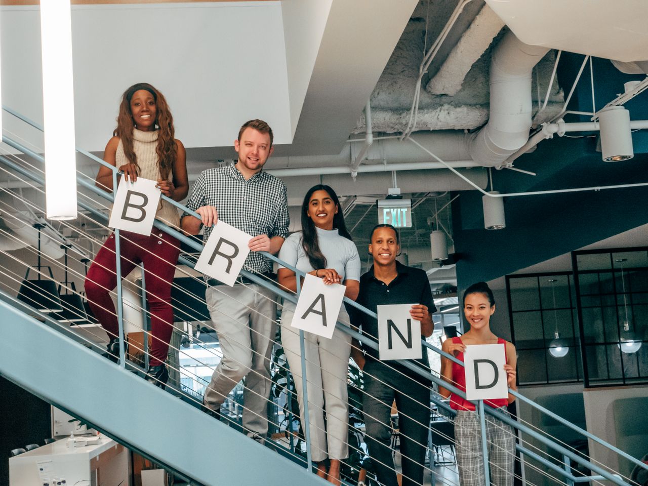 services-04 Diverse group of adults smiling and holding 'BRAND' signs on an office staircase.